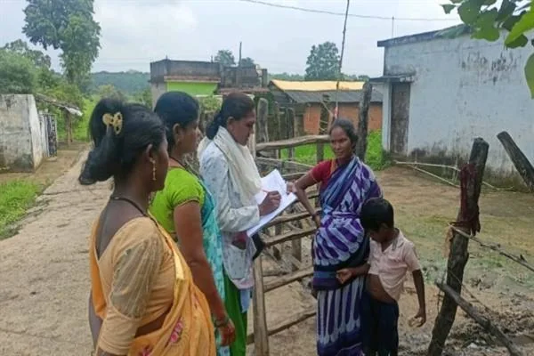 गांव में एक महिला अधिकारी कागज पर जानकारी लिखते हुए, पास खड़ी महिलाओं और एक बच्चे से बात कर रही हैं