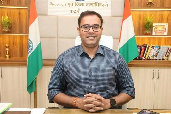 कार्यालय में कुर्सी पर बैठे चश्मा पहने अधिकारी, पीछे दोनों ओर भारतीय ध्वज और अलमारियां