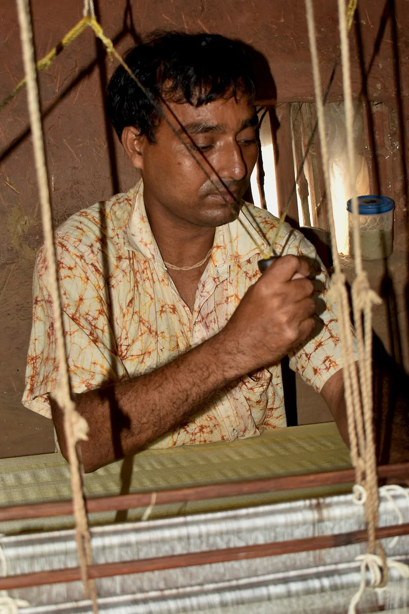 पश्चिम बंगाल में लकड़ी के हाथकरघे पर बैठकर कपड़ा बुनते एक बुनकर
